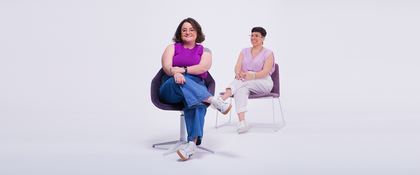 Na imagem, duas mulheres brancas e de cabelos curtos estão sentadas em cadeiras roxas em um espaço todo branco. Elas vestem roupas em tons de roxo e sorriem para a foto.