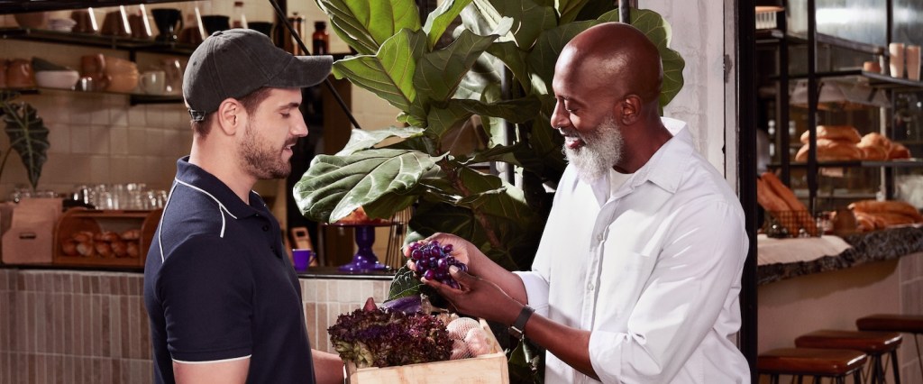 Foto de dois homens, um mais jovem branco, com boné, segurando uma caixa de madeira com alface, alho, uvas e outros alimentos dentro e outro negro, mais idoso, observando.