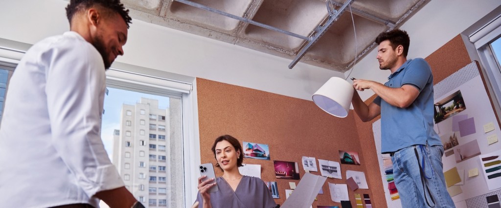 Foto de três pessoas (um mulher no meio e dois homens ao lado dela). Ela está segurando o celular numa mão e uma folha de papel na outra. Eles estão numa sala de trabalho, que tem um painel com informações pregadas e um abajur.