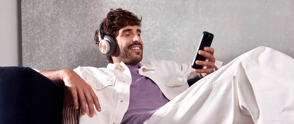 Homem branco está sentado num sofá azul escuro. Ele veste uma camiseta roxa, uma camisa e calça brancas. Ele tem barca e cabelos escuros. Usa um fone grande no ouvido, segura e olha para o celular e sorri.