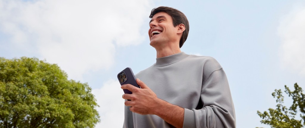 Imagem mostra homem branco, de cabelos lisos, sorrindo ao ar livre. O céu está azul. Ele veste moletom cinza.