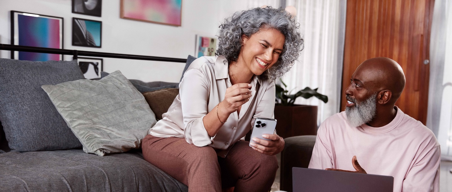 Imagem mostra uma mulher negra, com cabelos grisalhos sentada ao lado de um homem negro. Os dois estão sorrindo e olham o celular.