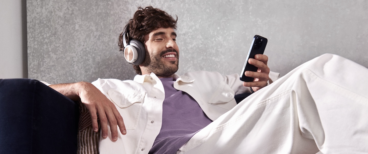 Imagem mostra um homem branco jovem, com barba e cabelos castanhos, sentado em um sofá, com celular na mão e fone no ouvido. Ele está olhando o celular e sorrindo. Ele veste camiseta roxa e jaqueta branca.