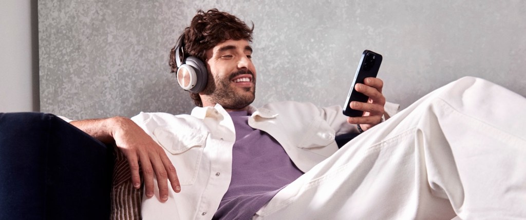 Imagem mostra um homem branco jovem, com barba e cabelos castanhos, sentado em um sofá, com celular na mão e fone no ouvido. Ele está olhando o celular e sorrindo. Ele veste camiseta roxa e jaqueta branca.