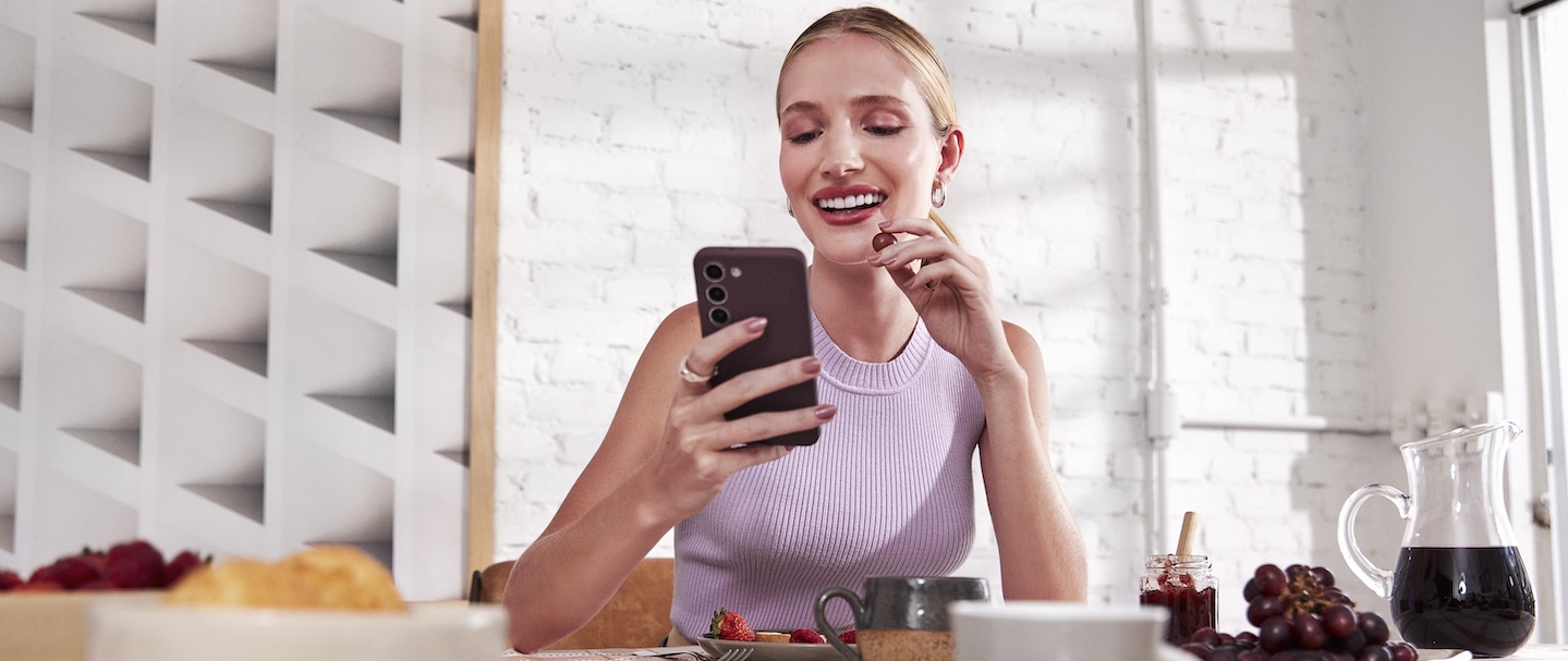 Imagem mostra uma mulher branca, loira, setada à mesa, comendo uma fruta e olhando para o celular. Ela veste uma regata roxa.