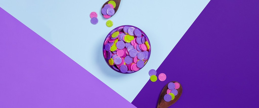 imagem com fundo roxo e azul mostra moedas coloridas, rosa, limão e roxas. E duas colheres de pau.