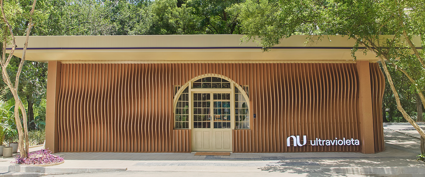 Casa Nubank Ultravioleta: tudo sobre o seu refúgio no Parque Ibirapuera. A imagem destaca a elegante fachada da casa, harmoniosamente integrada ao cenário natural do Parque Ibirapuera. Com um design moderno que utiliza linhas verticais de madeira, a estrutura se funde com a paisagem ao redor. O logotipo "Nu Ultravioleta" é visível no canto inferior direito, simbolizando sofisticação e modernidade. Predominam os tons terrosos, enriquecidos por uma linha na cor ultravioleta no teto que acrescenta um toque vibrante. A entrada é marcada por uma porta clássica com quadrados de vidro transparente, oferecendo charme atemporal. Um tapete bege à porta proporciona uma recepção acolhedora, equilibrando exclusividade e elegância.