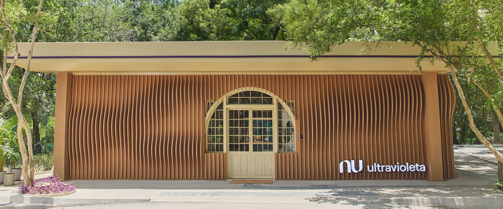 Casa Nubank Ultravioleta: tudo sobre o seu refúgio no Parque Ibirapuera. A imagem destaca a elegante fachada da casa, harmoniosamente integrada ao cenário natural do Parque Ibirapuera. Com um design moderno que utiliza linhas verticais de madeira, a estrutura se funde com a paisagem ao redor. O logotipo 