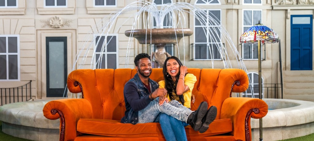 Foto mostra um homem negro, com calça e jaqueta jeans; e uma mulher asiática, de camiseta amarela e calça jeans sentados num sofá laranja em frente a uma fonte, que é um dos cenários da série friends.