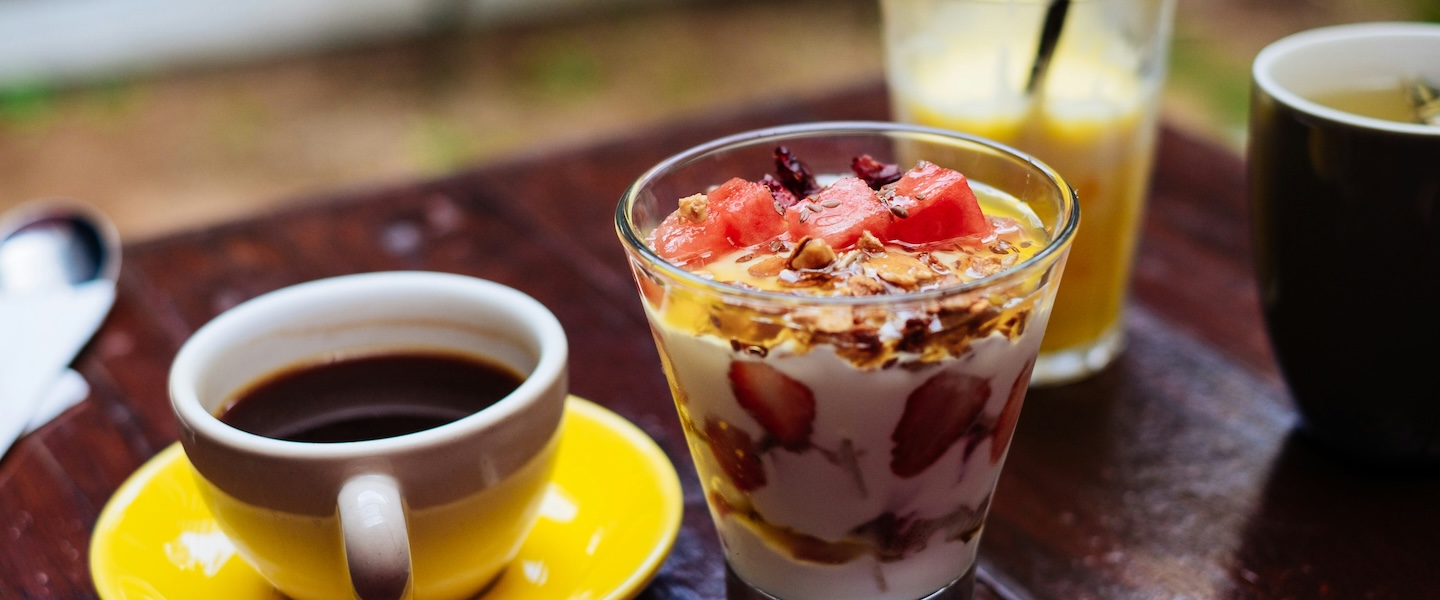 Foto de uma mesa de madeira. Em cima: uma xícara com café, um copo com iogurte e frutas (morango, melancia, granola), um copo de suco de laranja ao lado. Foto: Vince Schwidder