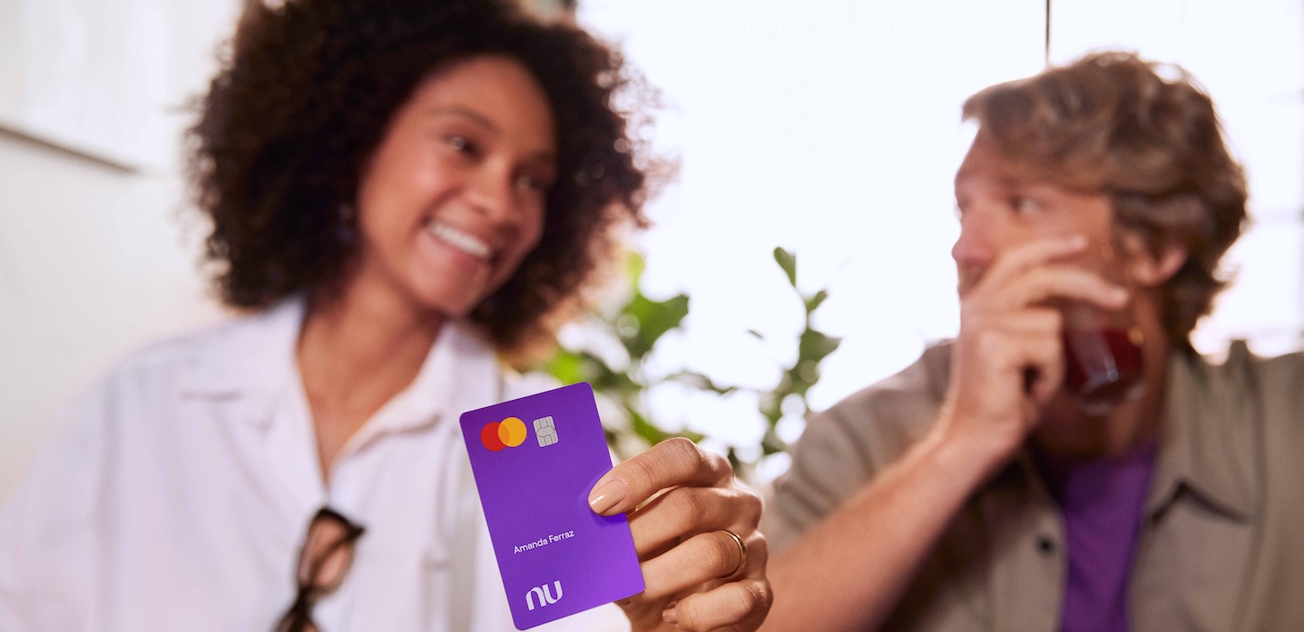 Foto mostra uma mulher negra, com cabelos curtos e cacheados, sorrindo, e segurando um cartão roxo do Nubank, que está em primeiro plano. Ao lado dela, um homem branco, com cabelos lisos, curtos e louros, olha para ela. Ele segura um copo.